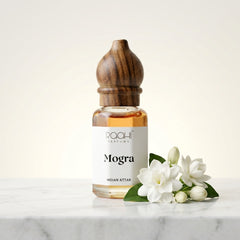 A clear glass bottle with a dark wood cap labeled mogra indian attar by roahi parfums handcrafted in kannauj
