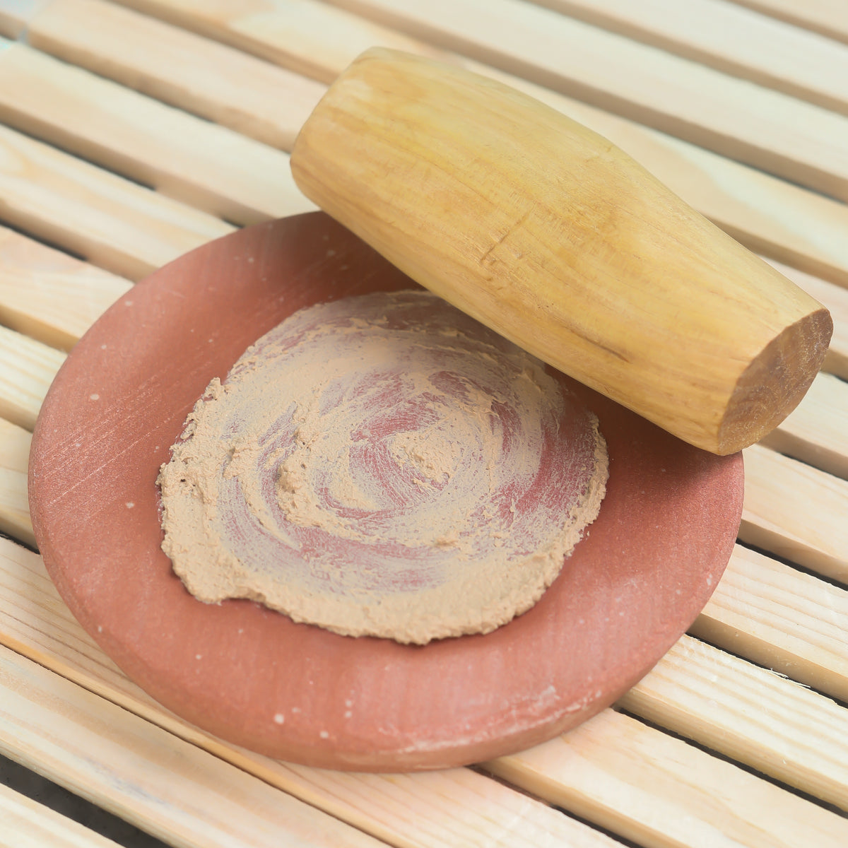 A light brown wooden pestle beside a terracotta mortar with beige and reddish sandalwood powder for rubbing use